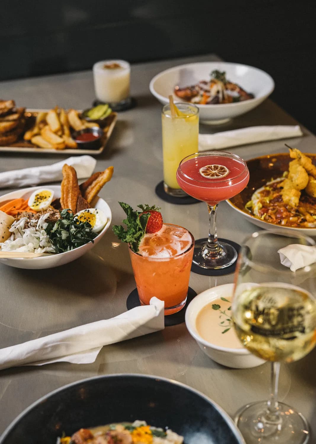 A table spread of Sweet Fish dishes and cocktails, including a ramen bowl, fries, and seafood.