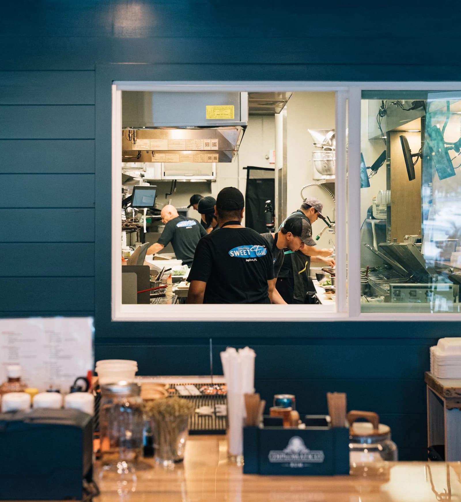Sweet Fish cooks in branded black tees working the open kitchen line, seen through the pass window above the service counter.